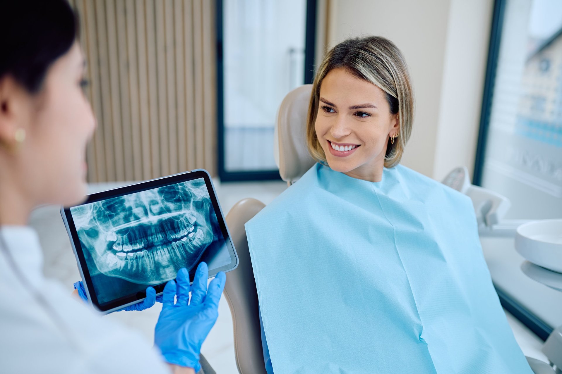 Dentist in blue gloves holding tablet with patient's panoramic dental x-ray while talking to happy female patient in clinic