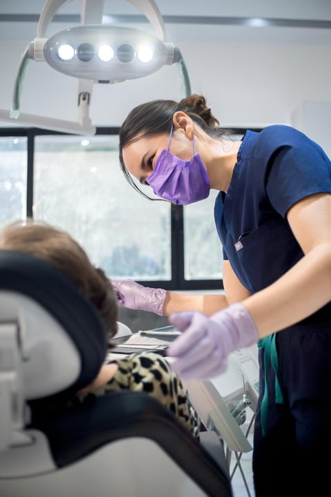 Dentist in blue scrubs and purple mask examining a patient under overhead dental light in clinic