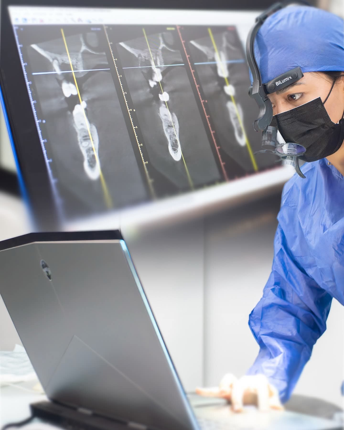 Medical professional in blue scrubs and mask viewing dental X-rays on a monitor while working at a laptop